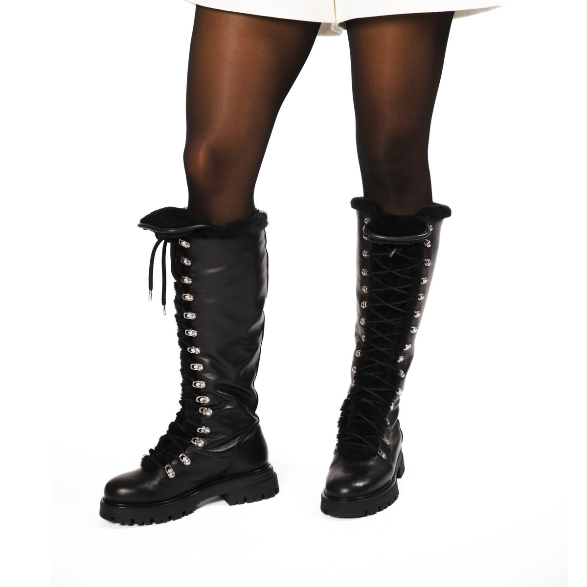 Scarlett Chase Aspen Knee-High Winter Shearling Boot in Black Leather with silver nickel eyelets and a black lace | Woman's legs showing 3/4 front view of boots dressed in black nylons and white short garment