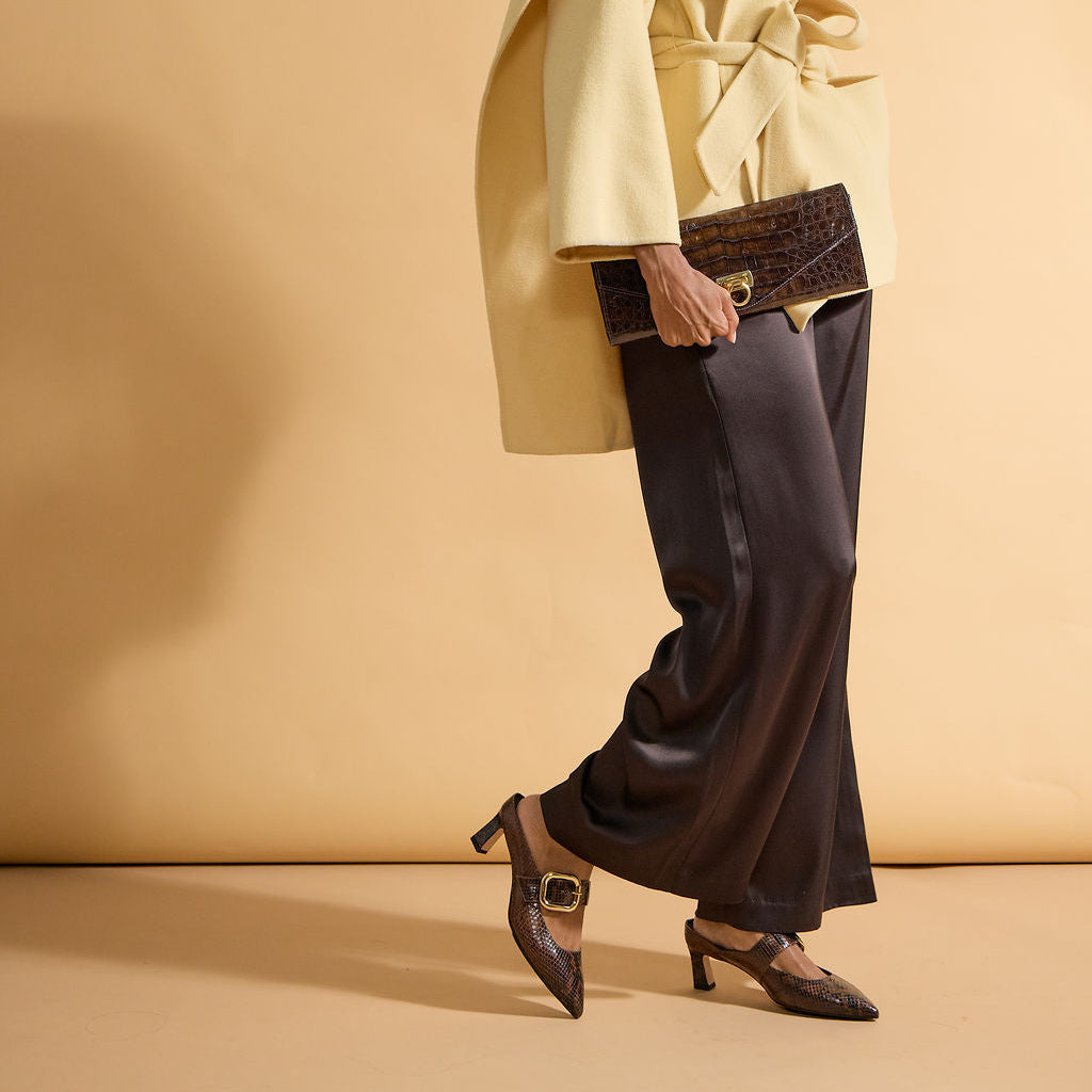 A model is shown from a side-back angle wearing a pale yellow belted cape jacket, dark brown wide-leg trousers, and dark brown pointed-toe heels, holding a dark brown crocodile-embossed clutch against a solid beige studio backdrop.