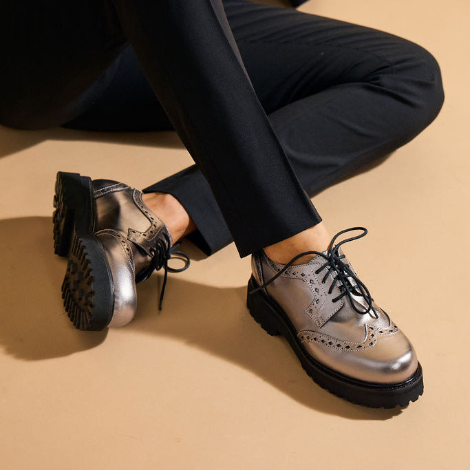 A person wearing shiny metallic silver lace-up oxford shoes with black soles, shown from the knees down while sitting on a beige floor.