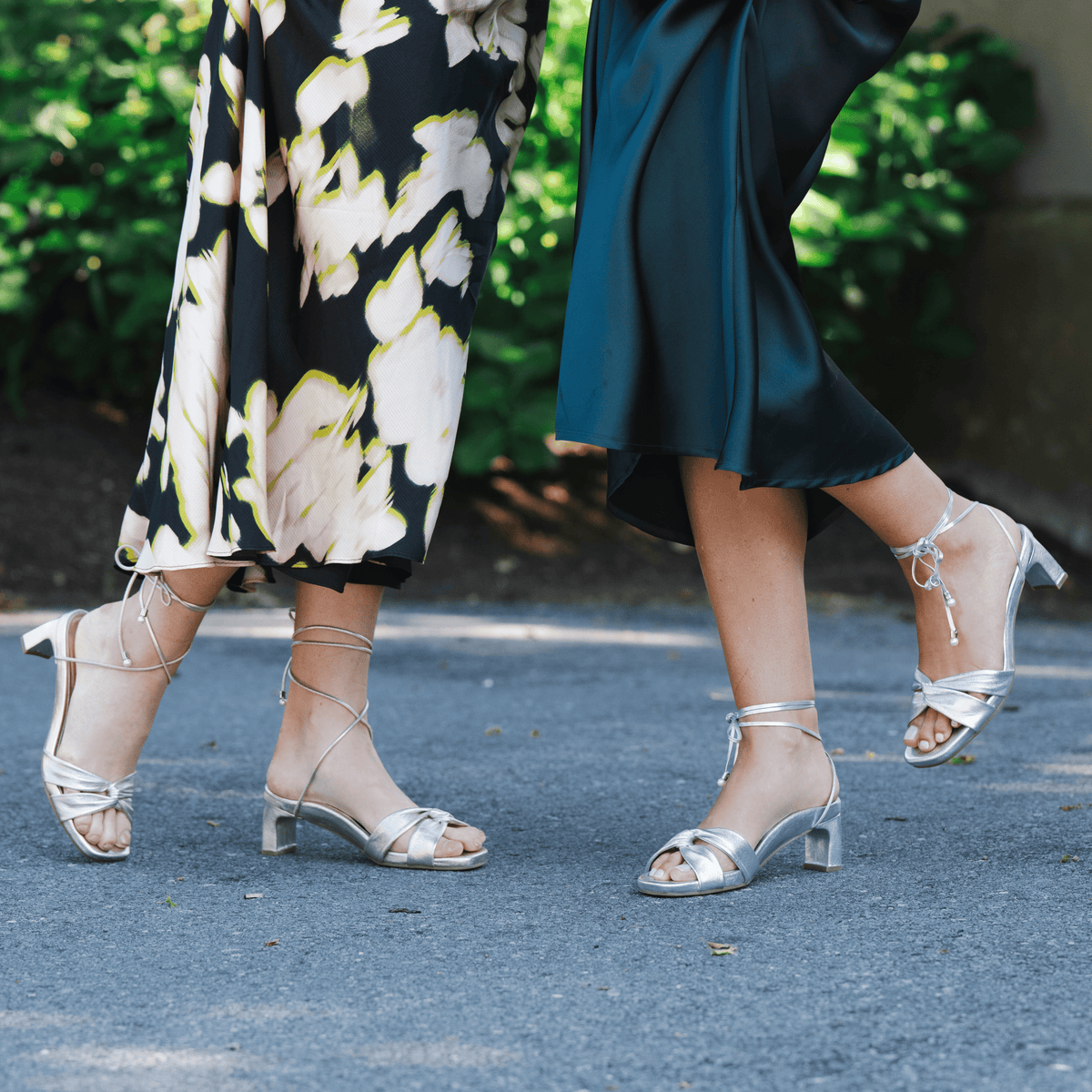 Scarlett Chase silver metallic and champagne napa leather Cali 45 dress sandal with wrap-around ties and a gathered front | 2 models side by side outside on driveway left model has one foot heel raised and profile view other model 3/4 view with back shoe off ground