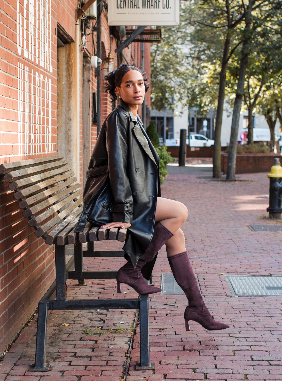 Scarlett Chase knee-high stretch wine suede Lola 75 boot with a block heel | Model on bench in city setting side view of boots one on rung of bench other hanging in air