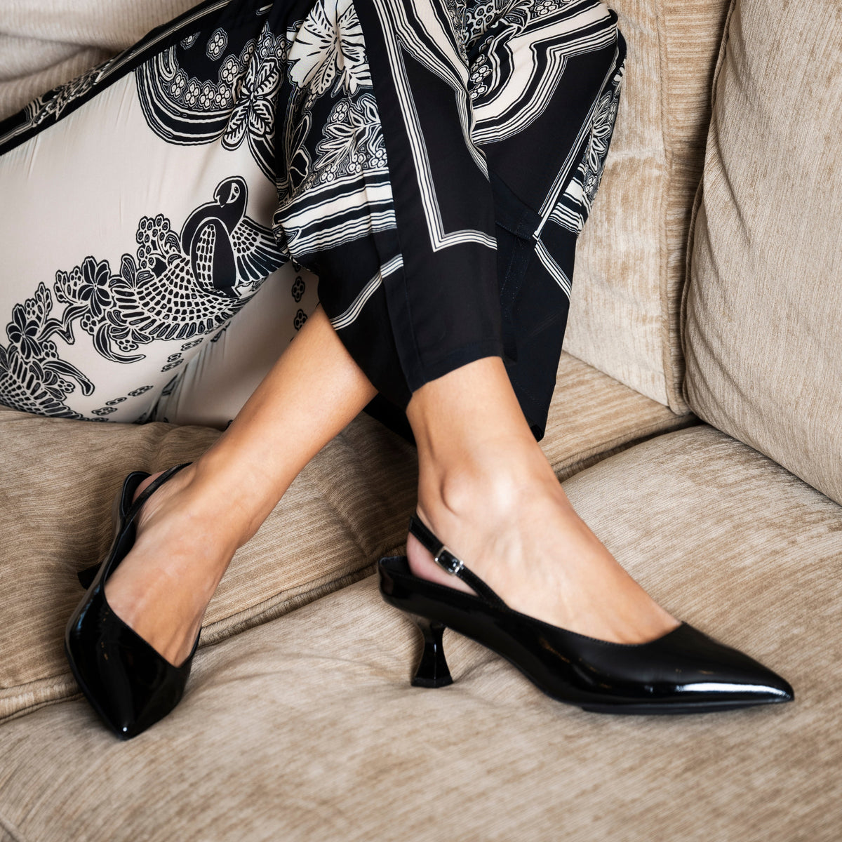 Scarlett Chase crinkle black patent Reese 55 Slingback with a sculpted heel, an adjustable backstrap buckle, and a toe accessory | On model bent knees with patterned pants on cream couch front side view of one shoe and side view of other