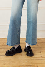 Person wearing blue jeans and black  patent Scarlett Chase Black Patent Arden Lug loafers on a wooden floor.