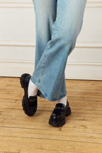 Person wearing light blue jeans with crossed legs wearing Scarlett Chase Arden Lug Loafers on a wooden floor.