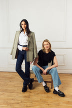 Scarlett Chase Ava Lug Penny Loafer in Black Leather with a lightweight sole and SC coin | Two women posing (one standing, one sitting) in a room with wooden floor and white walls. One wearing Scarlett Chase Arden Lug Loafers, the other wearing the Ava Lug Penny Loafer
