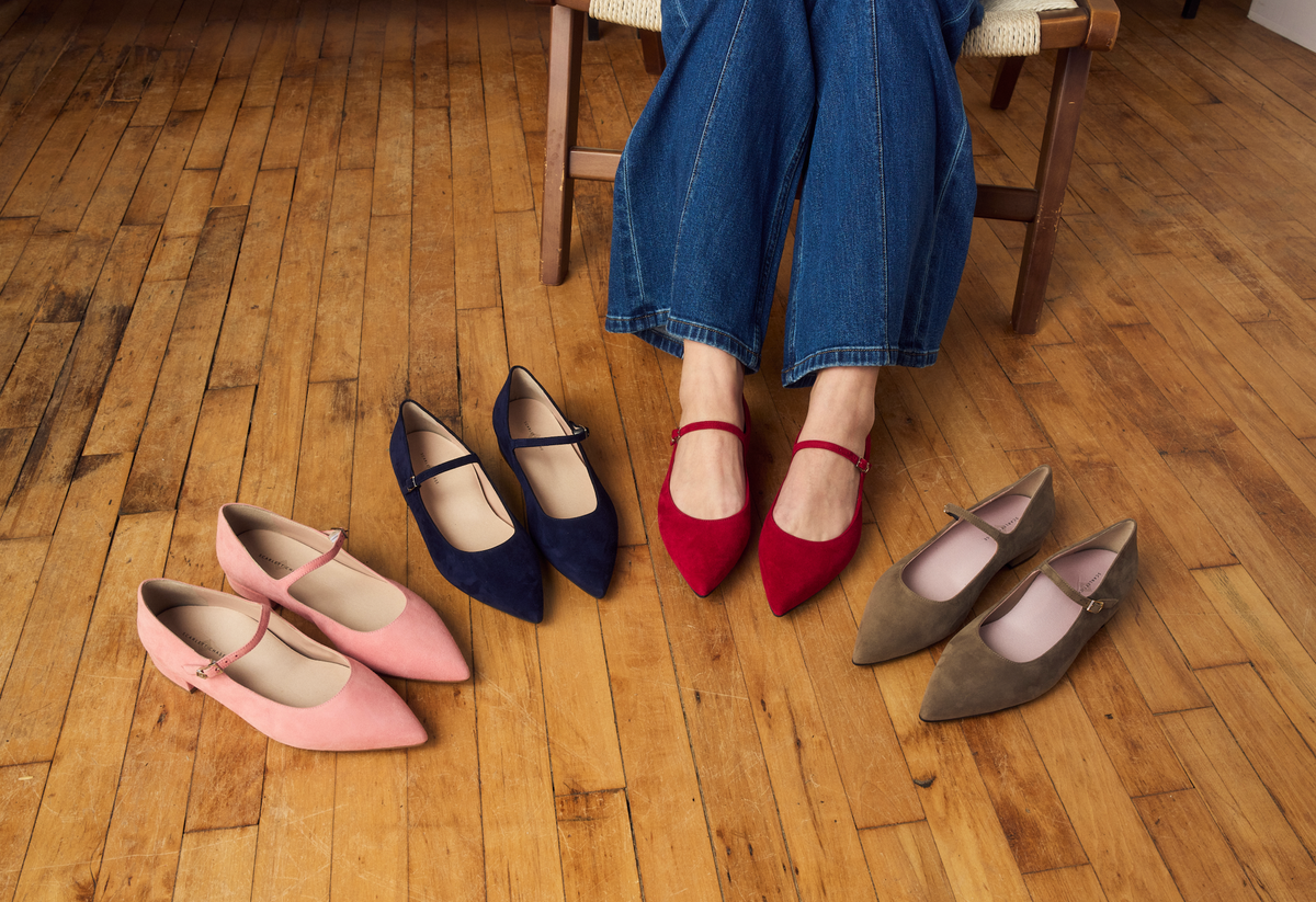 Woman sitting on a chair with four pairs of Scarlett Chase Belle Ballet Suede Mary-Jane Flats in front of her on a wooden floor.