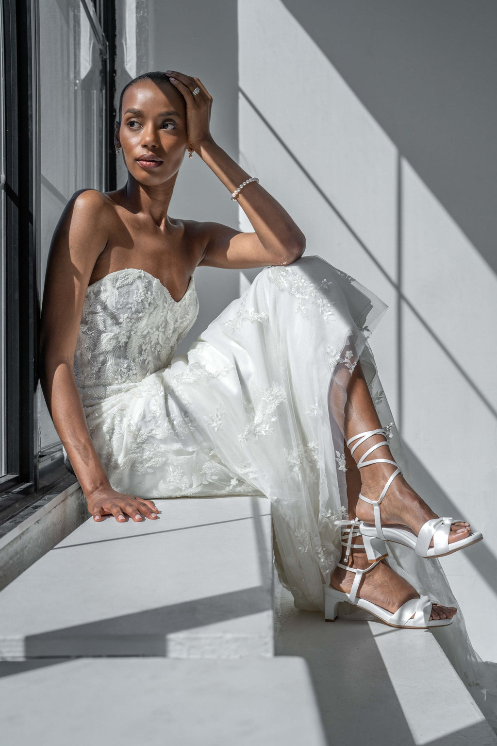 Scarlett Chase Cali 45 white satin dress sandal with wrap-around ties and a gathered front | Woman in a white wedding dress sitting by a window with sunlight casting shadows.