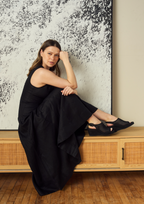Scarlett Chase Chelsea Wedge Sandal with a perforated leather upper and adjustable back strap | Woman in a black dress sitting on a wooden bench against a textured wall.
