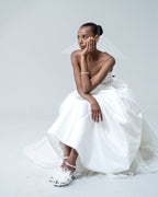 Scarlett Chase white satin Coco 45 dress sandal with minimalist straps and an adjustable backstrap and large clip-on Contessa white feather plumette shoe accessory | On seated bride 3/4 view with plumette  on pair facing left on light blue background