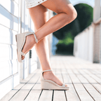 Scarlett Chase dune metallic napa leather Corsica Wedge Sandal with an adjustable back strap and handroped wedge bottom | On model feet one leg leaning against wall outside on deck one leg flat on ground side view 