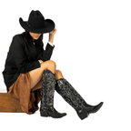 Scarlett Chase black leather Fiona cowboy bootie with decorative stitching and a side zipper and interchangeable knee-high gaiter with decorative stitching and clip-on straps | Pair with gaiters attached on model in cowboy attire with profile staggered view with one boot on the ground and the other with toe lifted on white background
