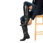 Scarlett Chase Flynn black leather riding boot with a side zipper and black / black leather Flynn interchangeable knee-high riding boot gaiter with classic upper band and clip-on straps | Side view of model sitting on stool wearing boot and knee-high gaiters with one foot in the air and hand on heel on white background