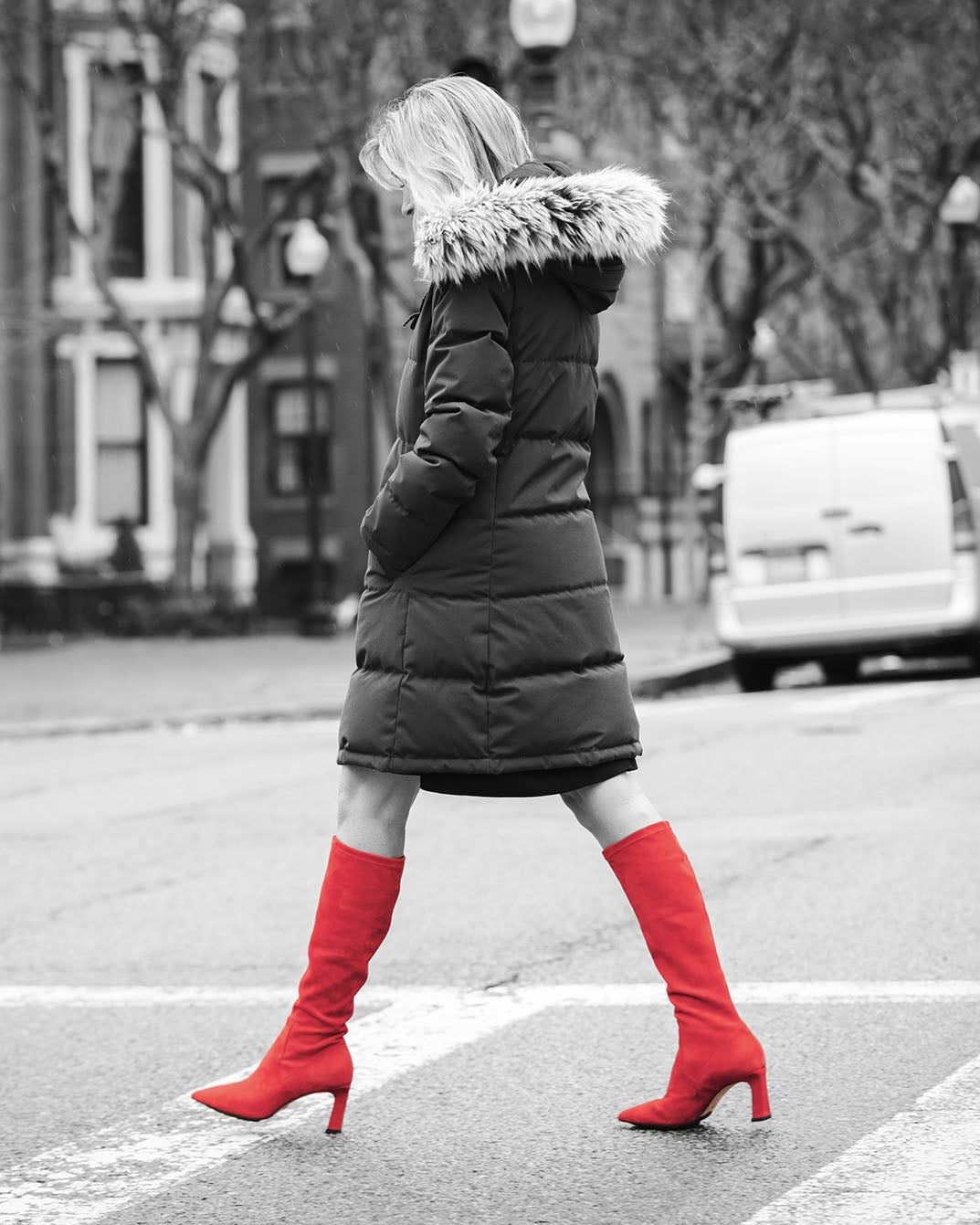 Person wearing a long dark coat with a fur-lined hood and bright red boots on a city street.