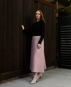 Scarlett Chase knee-high stretch rosa suede Lola 75 boot with a block heel | On feet 3/4 view of 1 shoe other heel view model in long pink skirt on white floor with wood background outside