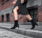Scarlett Chase ankle stretch black suede Portia 75 bootie with a block heel | Profile view of model jumping off curb onto cobblestones with city background left food in air right foot on toe with heel in air