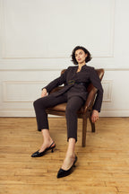 Scarlett Chase crinkle black patent Reese 55 Slingback with a sculpted heel, an adjustable backstrap buckle, and a toe accessory | On model sitting in chair in room with wooden floor and white backdrop 3/4 view of shoes