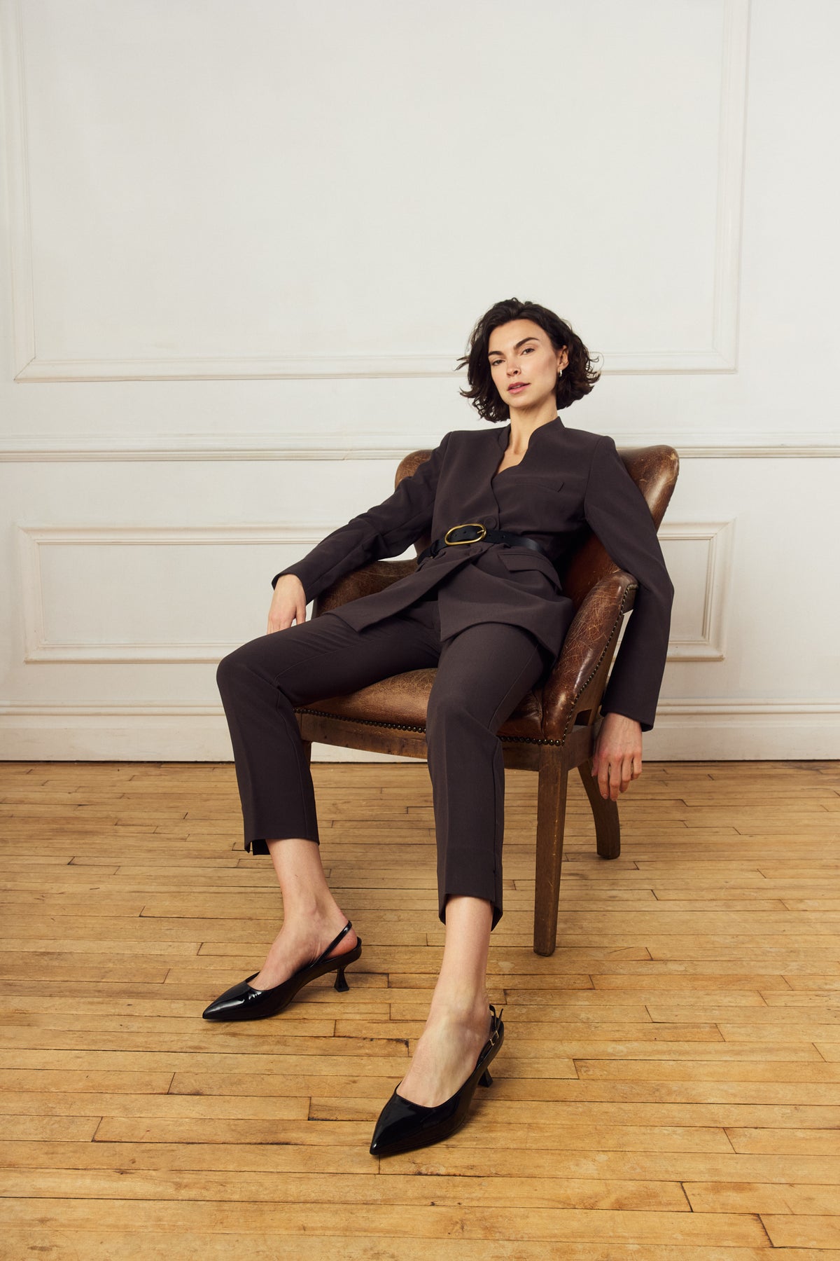Scarlett Chase crinkle black patent Reese 55 Slingback with a sculpted heel, an adjustable backstrap buckle, and a toe accessory | On model sitting in chair in room with wooden floor and white backdrop 3/4 view of shoes