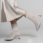 Scarlett Chase ankle stretch taupe suede Maya 55 bootie with a block heel  |  On model long white skirt walking with back leg bent shoe in air side view of both on white background