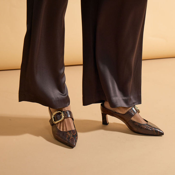 A person wearing dark brown satin wide-leg trousers and dark brown pointed-toe slingback heels with a snakeskin pattern and gold buckle detail, standing against a warm beige studio backdrop.