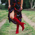 Scarlett Chase knee-high stretch red suede Lola 75 boot with a block heel | On model in dark colored velvet long dress on grass outside profile view of right boot in air and left front on ground
