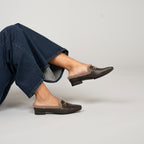 Scarlett Chase Weston Brown Leather and Camel Shearling Mule with an SC accessory | On foot of a model wearing jeans and lying down while lifting one foot on a grey background