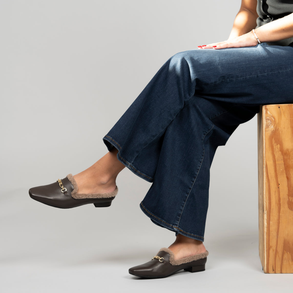 Scarlett Chase Weston Brown Leather and Camel Shearling Mule with an SC accessory | On foot of a model wearing jeans while sitting down on a wooden block cross legged on a grey background