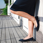 Scarlett Chase navy / silver italian suede Brigid ballet flat with a contrast heel and V-cut upper | Side profile on feet with model up against wall one shoe on wall and other on decking outside scenery navy skirt