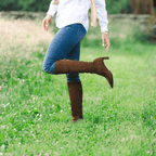 Clara 55 Scarlett Chase knee-high chocolate stretch suede Clara 55 boot with a block heel | On feet in tall grass one foot covered other foot lifted in air with sideview of boot country background