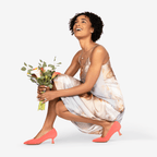 Scarlett Chase coral suede Gala 55 Pump with a rounded upper and sculpted heel | On model kneeling down with flowers tone foot ahead of the other back foot heel is off the ground on white background