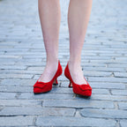 Scarlett Chase Rouge suede Greta 55 Pump with a rounded upper, sculpted heel, and red toe accessory | 3/4 of each side on feet with bare legs on black brick surface outside 