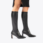 Scarlett Chase knee-high stretch onyx vegan leather Lola 75 boot with a block heel | On model black dress staggered stance profile view on white background
