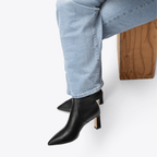 Scarlett Chase ankle stretch black onyx vegan leather Portia 75 bootie with a block heel |On model sitting on wood with white background 3/4 view of one other shoe toe and heel