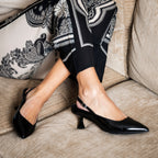 Scarlett Chase crinkle black patent Reese 55 Slingback with a sculpted heel, an adjustable backstrap buckle, and a toe accessory | On model bent knees with patterned pants on cream couch front side view of one shoe and side view of other 