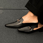 Scarlett Chase Weston Mule in Black Napa and an SC gold accessory | Side view on feet on Granite floor
