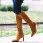 Scarlett Chase knee-high stretch camel suede Lola 75 boot with a block heel | On model with jeans tucked in staggered footing walking with back leg in air on white surface with greenery outdoors